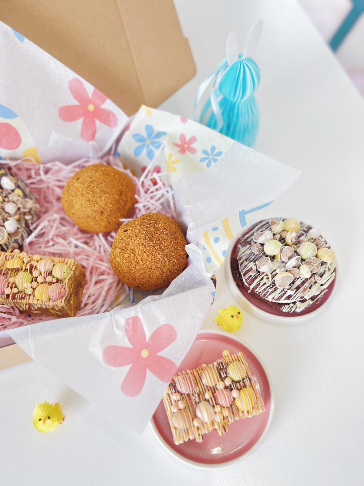An open box of Easter baked gifts in pretty patterned paper. Two Chocolate Scotch eggs, cookies and mini egg topped blondies for Easter. 