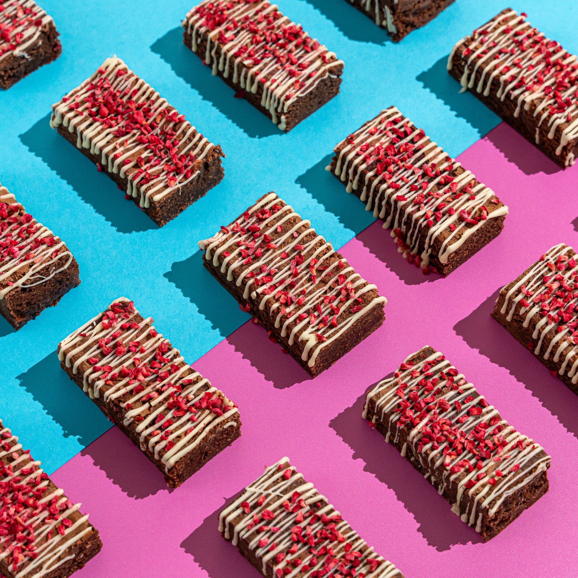 Fudgy raspberry and white chocolate brownies topped in white chocolate and freeze dried raspberry pieces, on a pale blue and pink background
